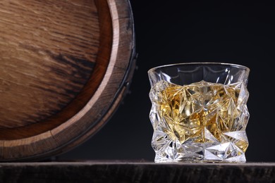 Whiskey with ice cubes in glass and barrel on wooden table against black background, closeup. Space for text