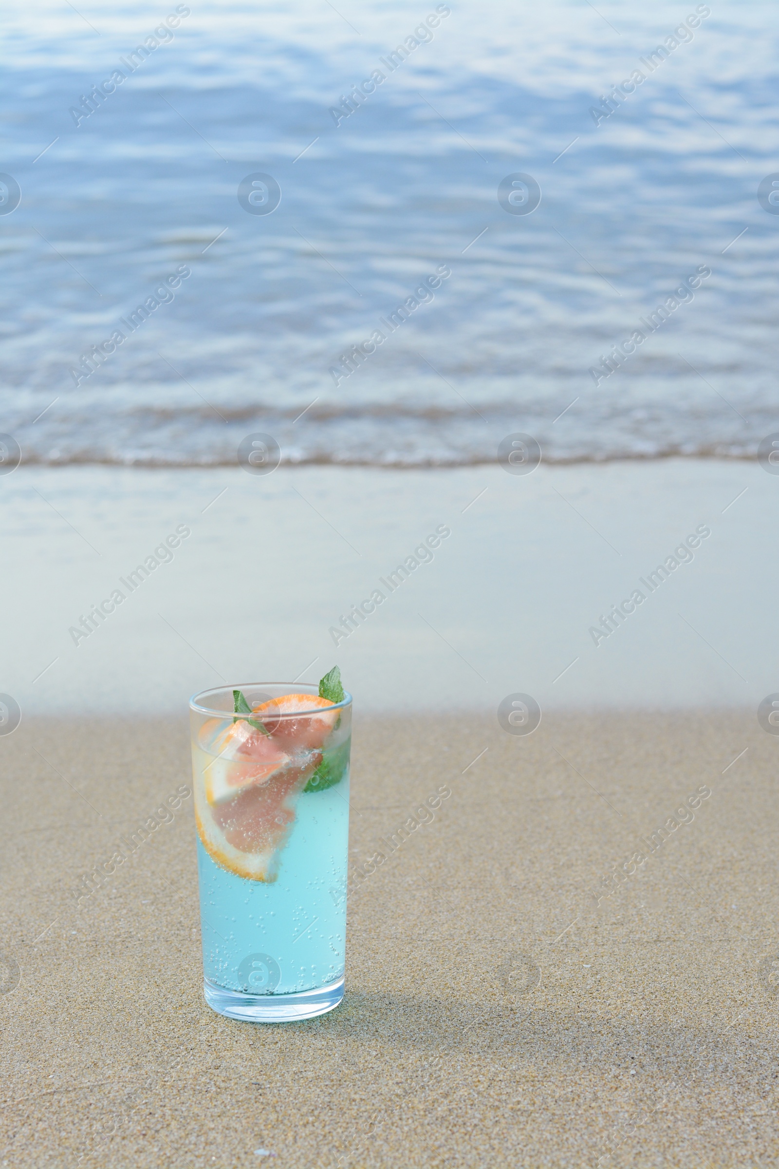 Photo of Glass of refreshing drink with grapefruit and mint on sandy beach near sea, space for text