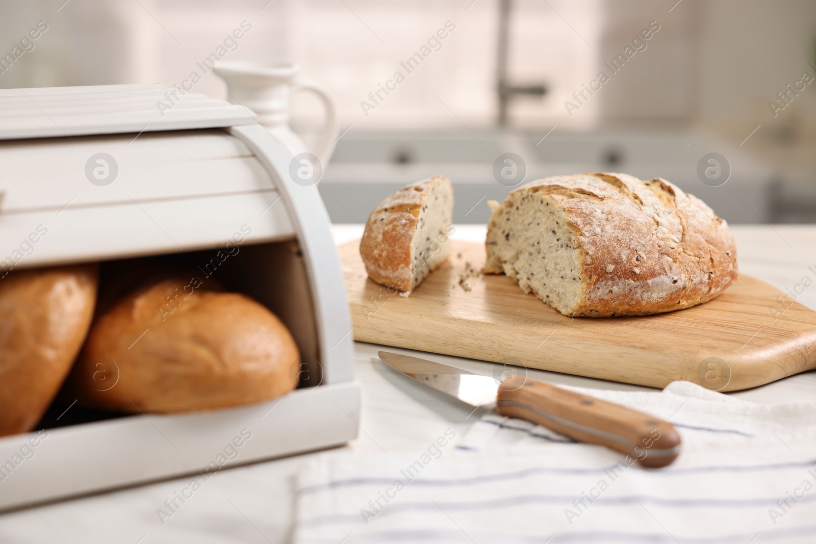 Photo of Wooden bread basket with freshly baked loaves and knife on white marble table in kitchen