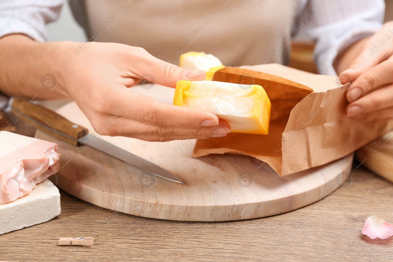 Photo of Woman putting natural handmade soap into paper bag at wooden table, closeup