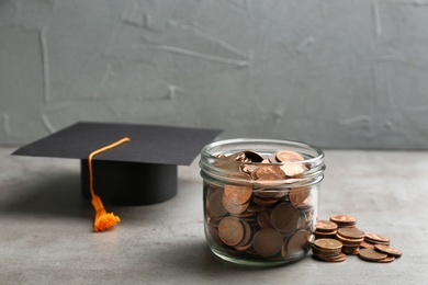 Glass jar, coins and graduation hat on grey table. Space for text