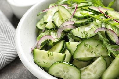 Photo of Delicious cucumber salad with onion and arugula in bowl on grey table, closeup