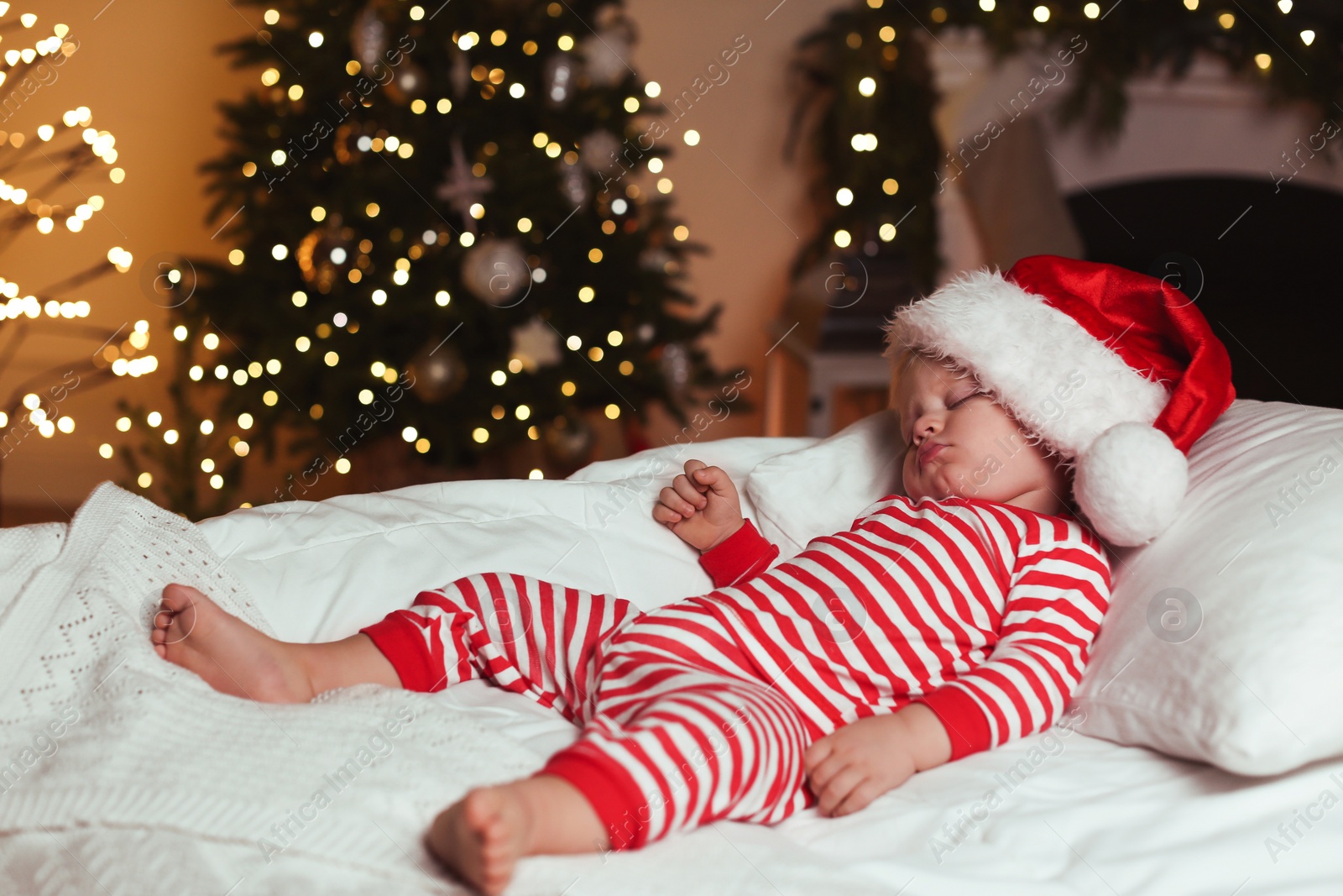 Photo of Baby in Christmas pajamas and Santa hat sleeping on bed indoors. Space for text