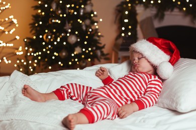 Photo of Baby in Christmas pajamas and Santa hat sleeping on bed indoors. Space for text