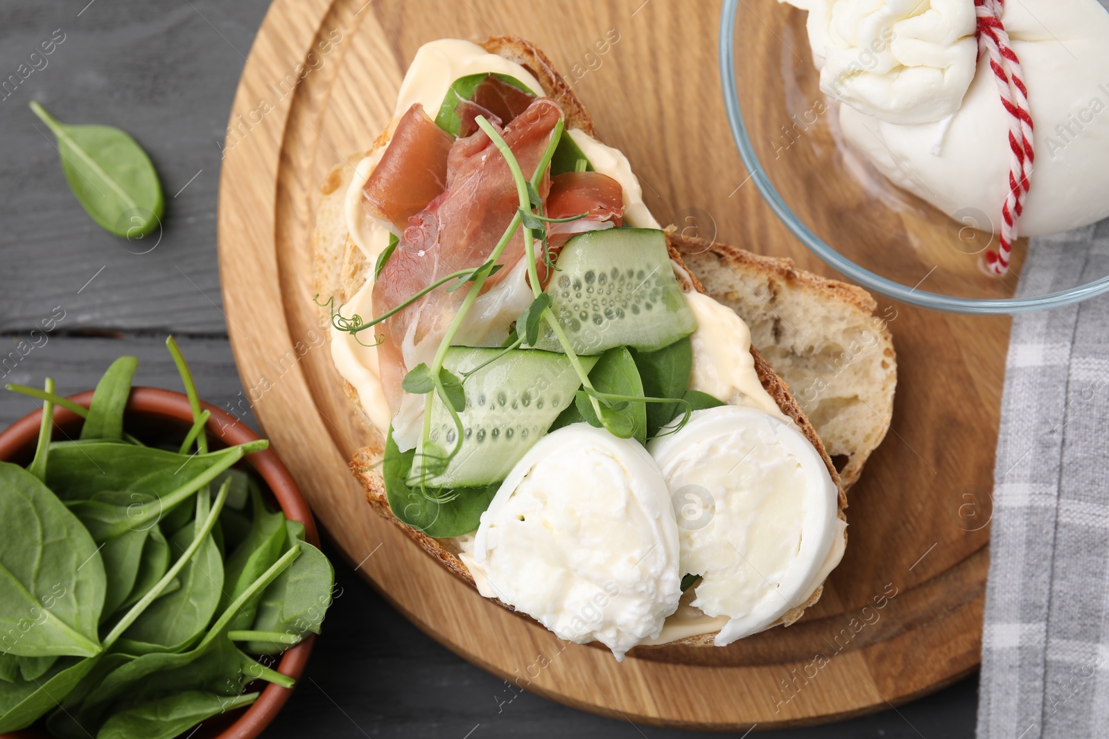 Photo of Tasty sandwich with burrata cheese, prosciutto, cucumber and ingredients on grey wooden table, flat lay