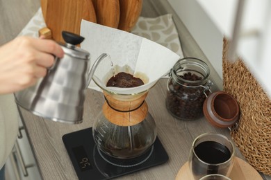 Woman pouring hot water into glass chemex coffeemaker with paper filter and coffee at countertop in kitchen, above view