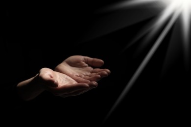 Image of Christian woman stretching hands towards holy light in darkness, closeup. Prayer and belief