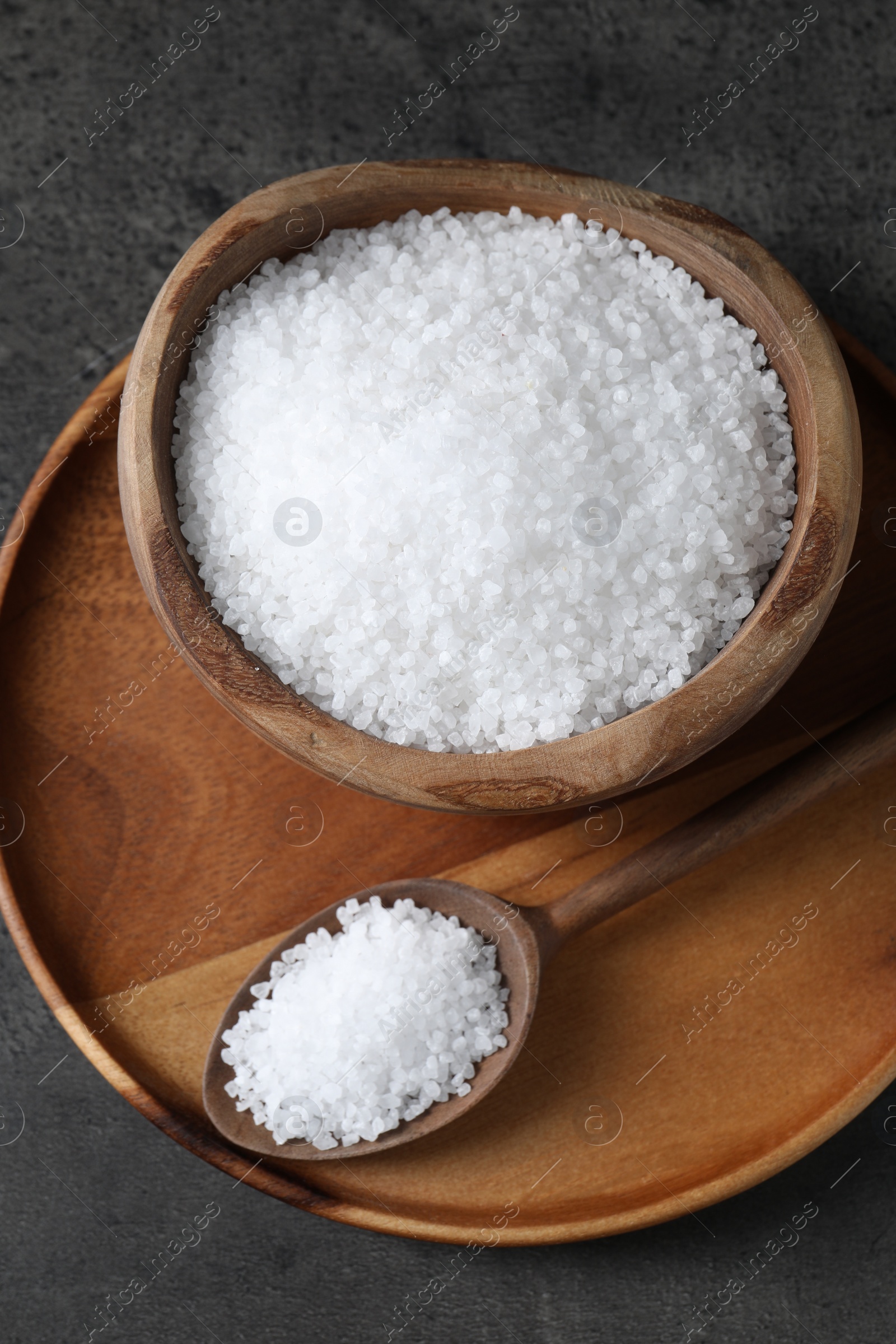 Photo of Natural salt in wooden bowl and spoon on dark grey table, top view