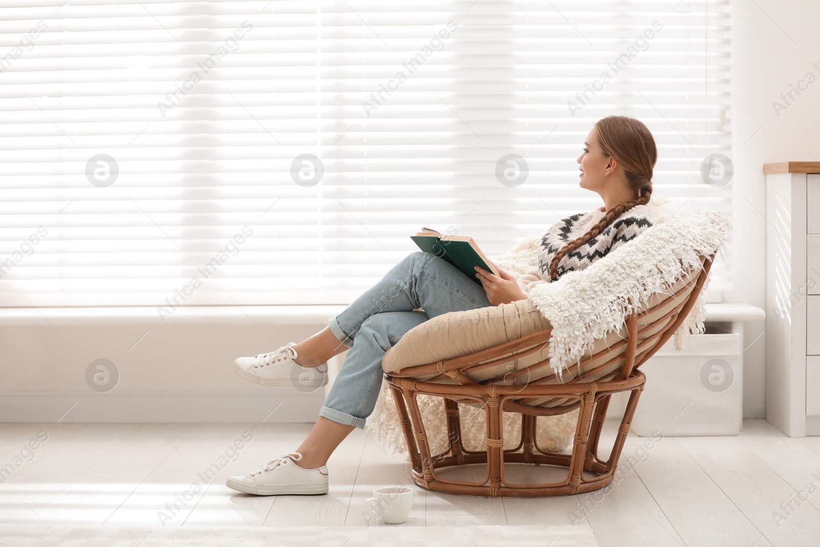 Photo of Young woman reading book in papasan chair near window at home