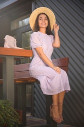 Photo of Beautiful young woman in stylish violet dress and straw hat sitting on bench outdoors