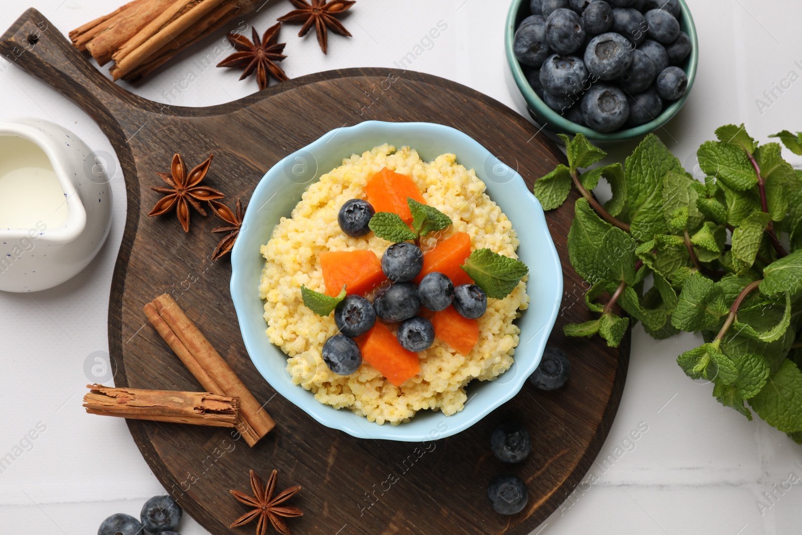 Photo of Tasty millet porridge with blueberries, pumpkin and mint in bowl on white tiled table, flat lay