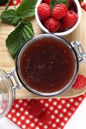 Photo of Jar of delicious raspberry jam, fresh berries and green leaves on white table, top view