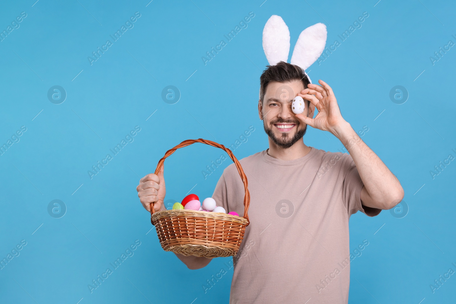 Photo of Happy man in bunny ears headband holding wicker basket with painted Easter eggs on turquoise background. Space for text