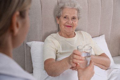 Photo of Young caregiver giving water to senior woman in bedroom. Home health care service