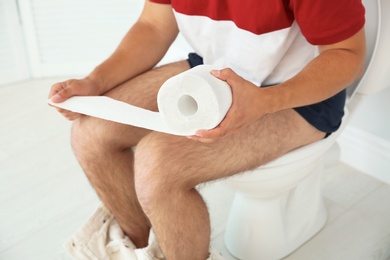 Photo of Young man with bath tissue sitting on toilet bowl at home