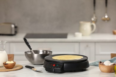 Cooking delicious crepe on electric pancake maker on white marble table in kitchen, space for text