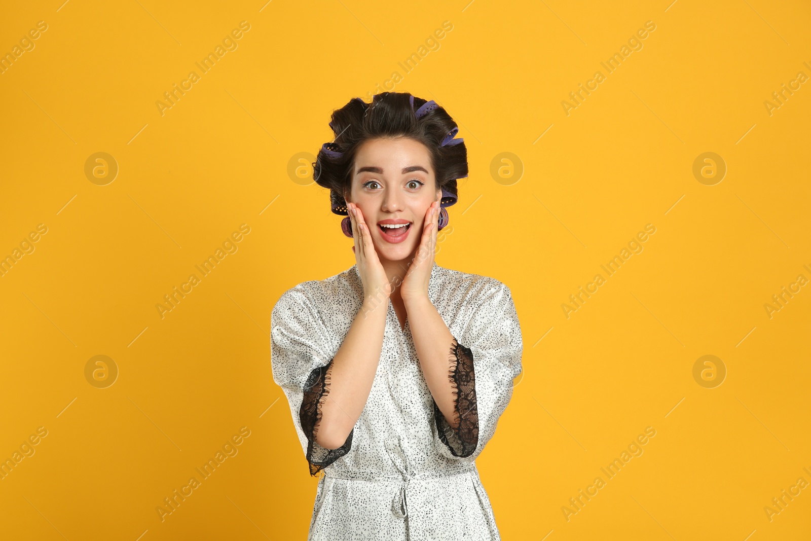 Photo of Emotional young woman in silk bathrobe with hair curlers on orange background