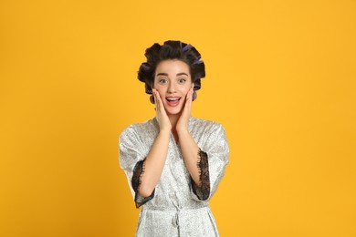 Emotional young woman in silk bathrobe with hair curlers on orange background