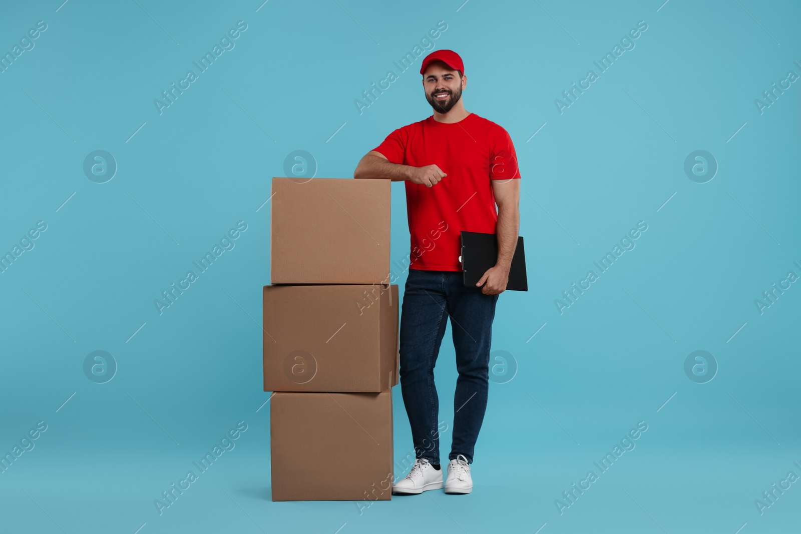 Photo of Happy courier with clipboard and stack of parcels on light blue background