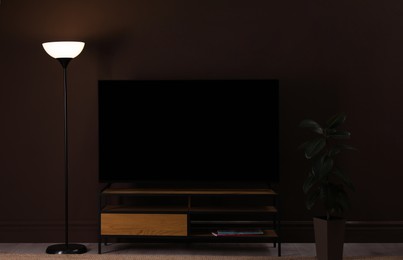 Photo of Modern TV on cabinet, floor lamp and houseplant near brown wall in room. Interior design