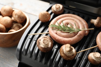 Photo of Electric grill with homemade sausage, rosemary and mushrooms on rustic wooden table, closeup