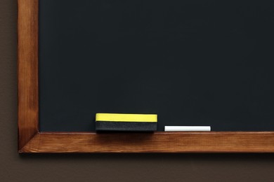 Photo of Clean blackboard with chalk and duster hanging on brown wall