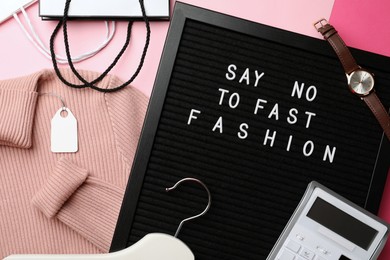 Black letter board with phrase SAY NO TO FAST FASHION, clothes, wristwatch, shopping bags, hanger and calculator on pink background, flat lay