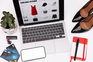 Photo of Online store website on laptop screen. Computer, smartphone, stationery, credit cards, women's shoes and lipstick on white background, above view