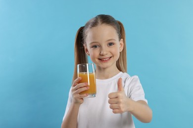 Cute little girl with fresh juice showing thumbs up on light blue background