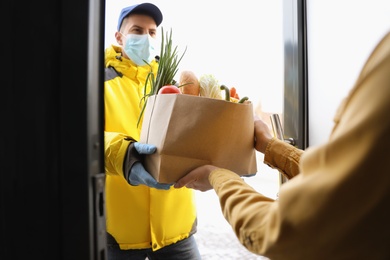 Photo of Courier in medical mask giving paper bag with groceries to woman at doorway. Delivery service during Covid-19 quarantine
