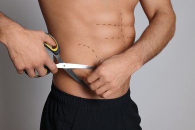 Photo of Fit man with scissors and marks on body against grey background, closeup. Weight loss surgery