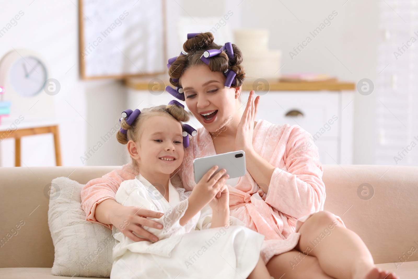 Photo of Happy mother and daughter with curlers and mobile phone on sofa at home