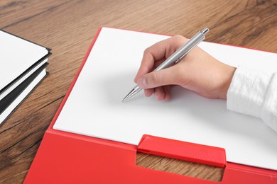 Photo of Woman writing on sheet of paper in red folder at wooden table, closeup