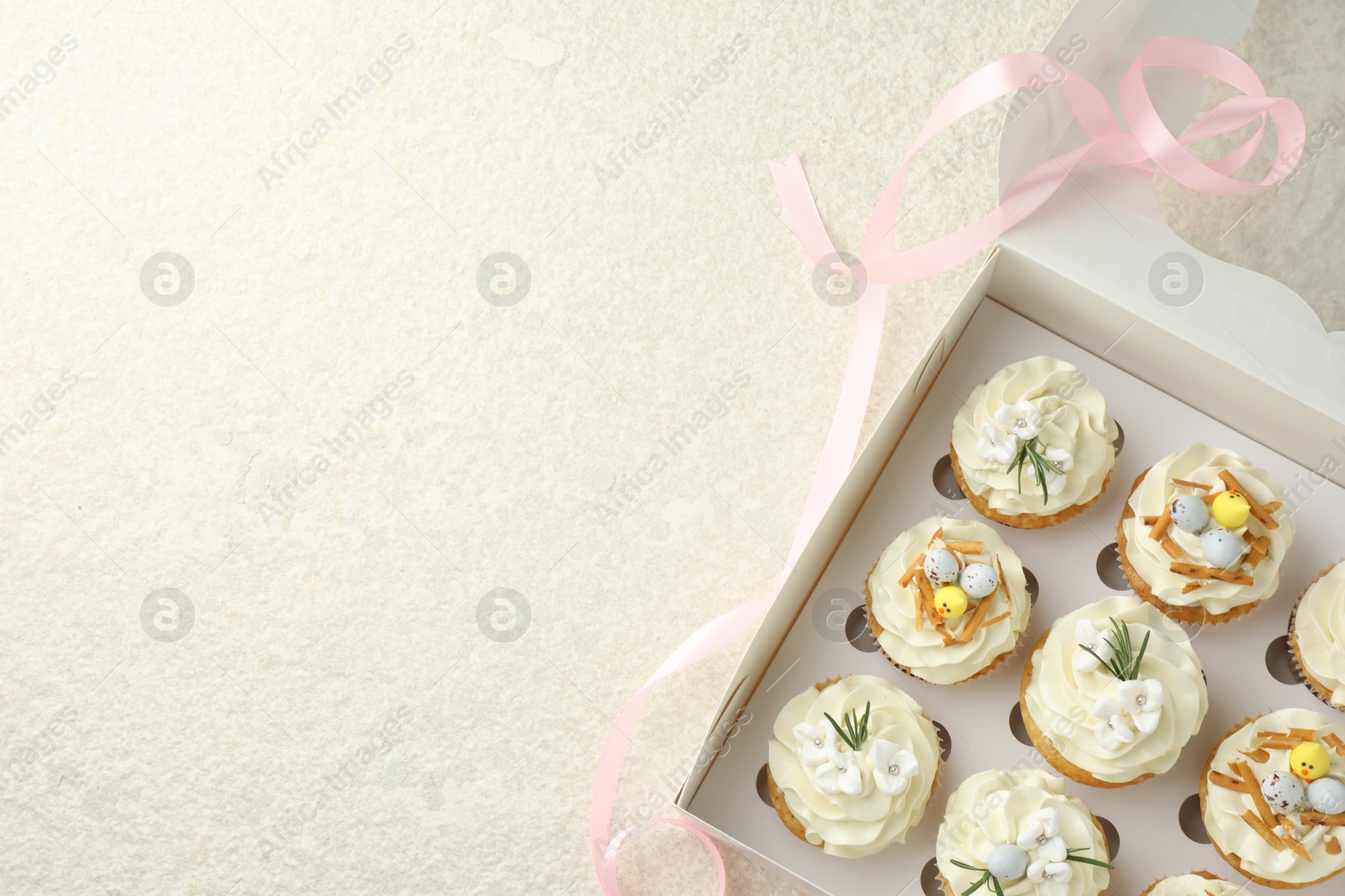 Photo of Tasty Easter cupcakes with vanilla cream in box and pink ribbon on beige table, flat lay. Space for text