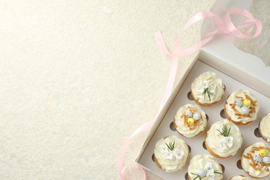 Photo of Tasty Easter cupcakes with vanilla cream in box and pink ribbon on beige table, flat lay. Space for text