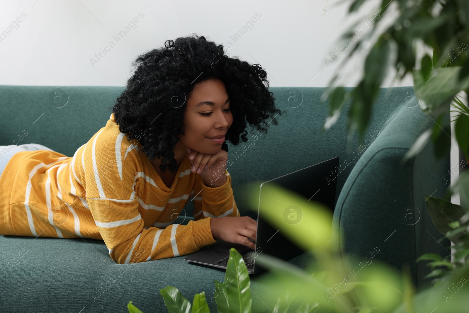 Photo of Relaxing atmosphere. Woman with laptop lying on sofa near houseplants indoors