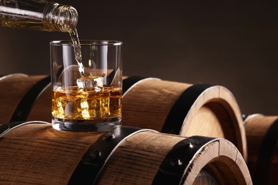 Photo of Pouring whiskey from bottle into glass on wooden barrel against dark background, closeup