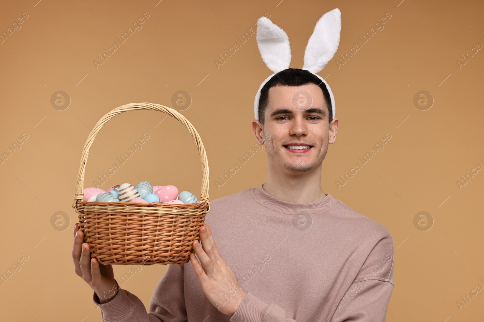 Photo of Easter celebration. Handsome young man with bunny ears holding basket of painted eggs on beige background