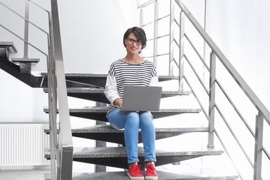 Photo of Young woman with modern laptop sitting on stairs indoors