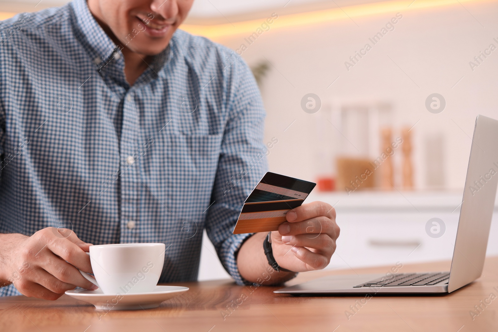 Photo of Man using laptop and credit cards for online payment at table in kitchen, closeup
