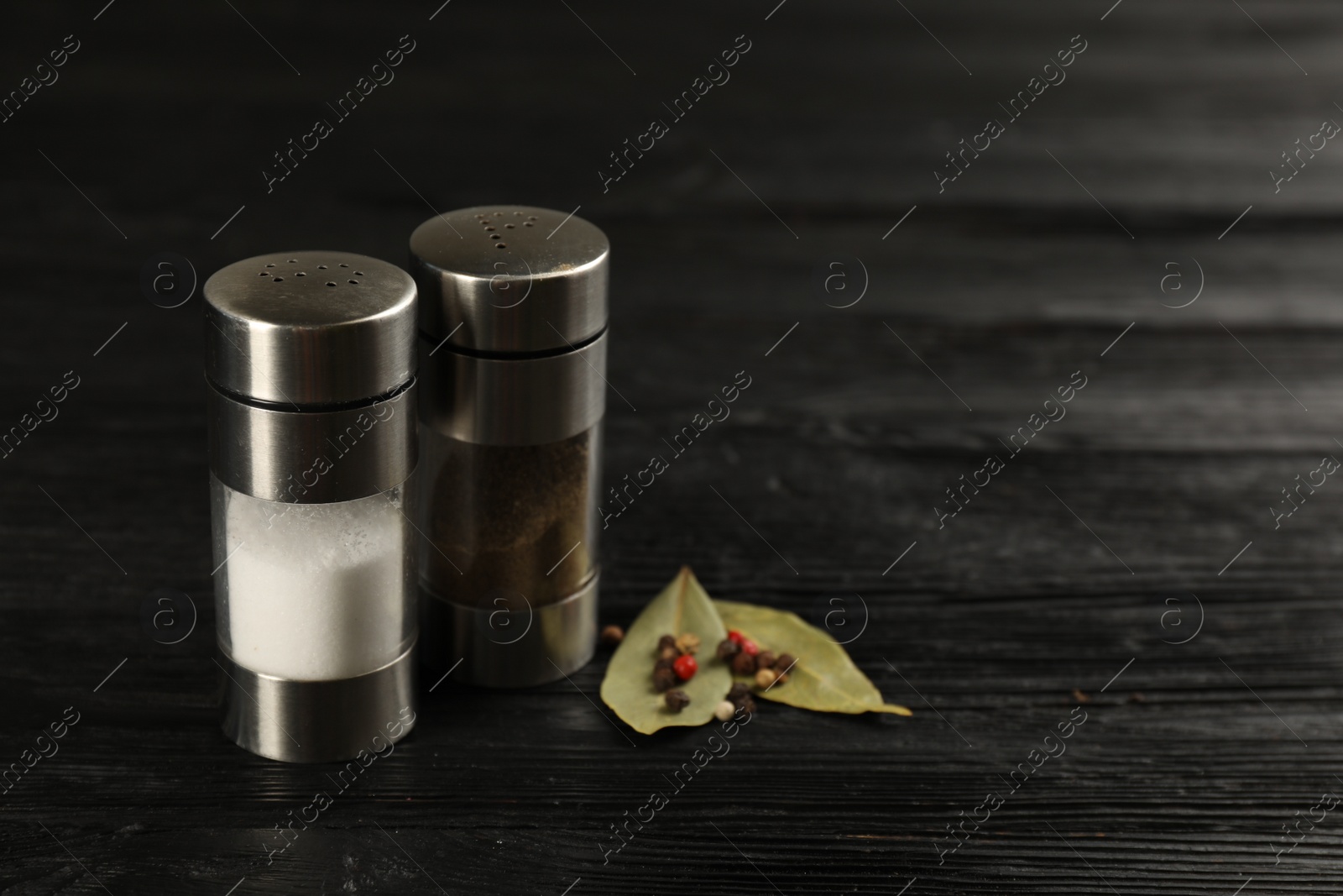 Photo of Salt and pepper shakers with bay leaves on black wooden table, closeup. Space for text