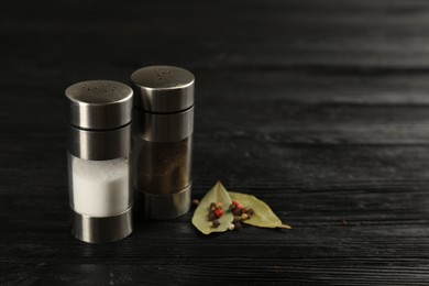 Photo of Salt and pepper shakers with bay leaves on black wooden table, closeup. Space for text