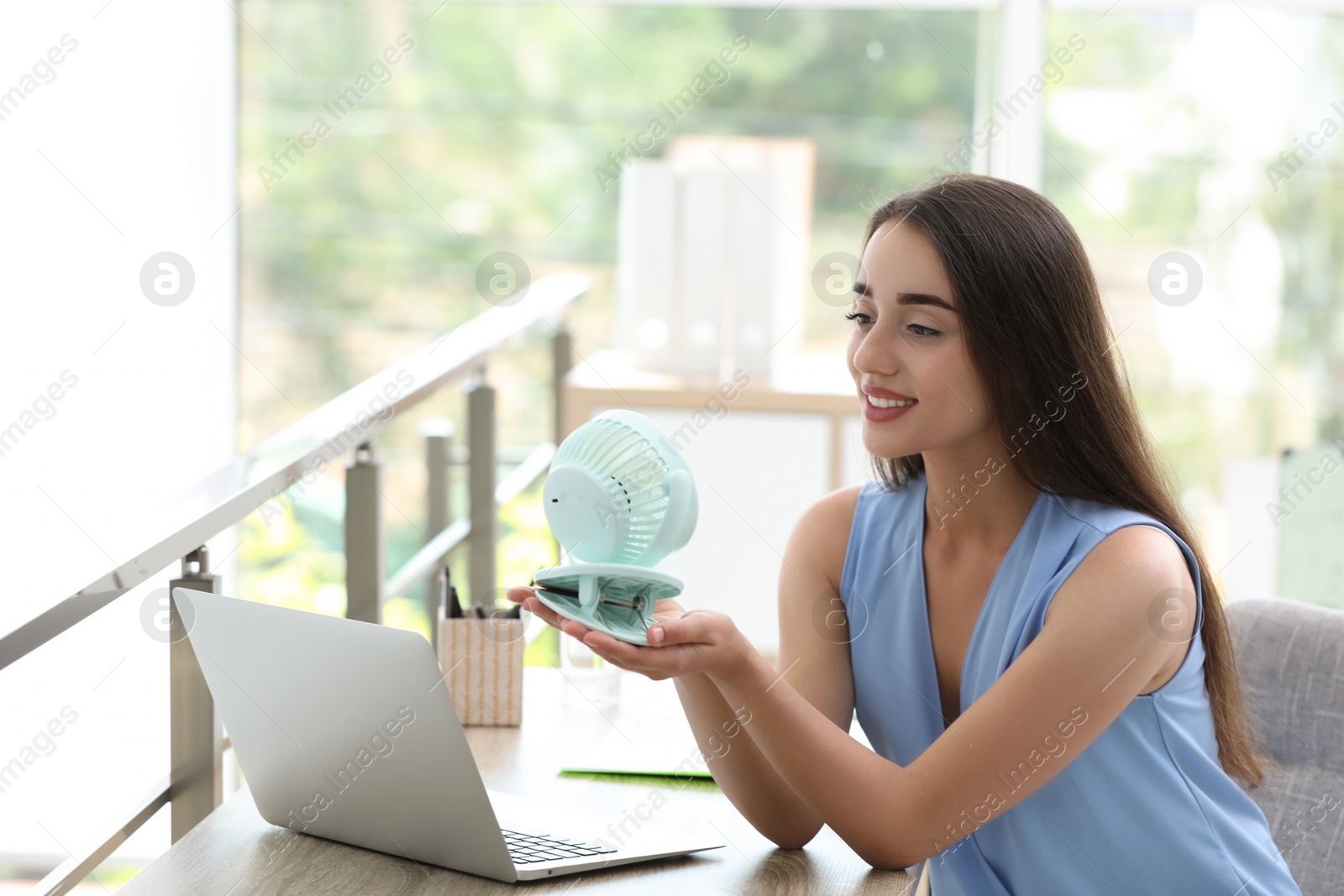 Photo of Young woman enjoying air flow from portable fan at workplace. Summer heat