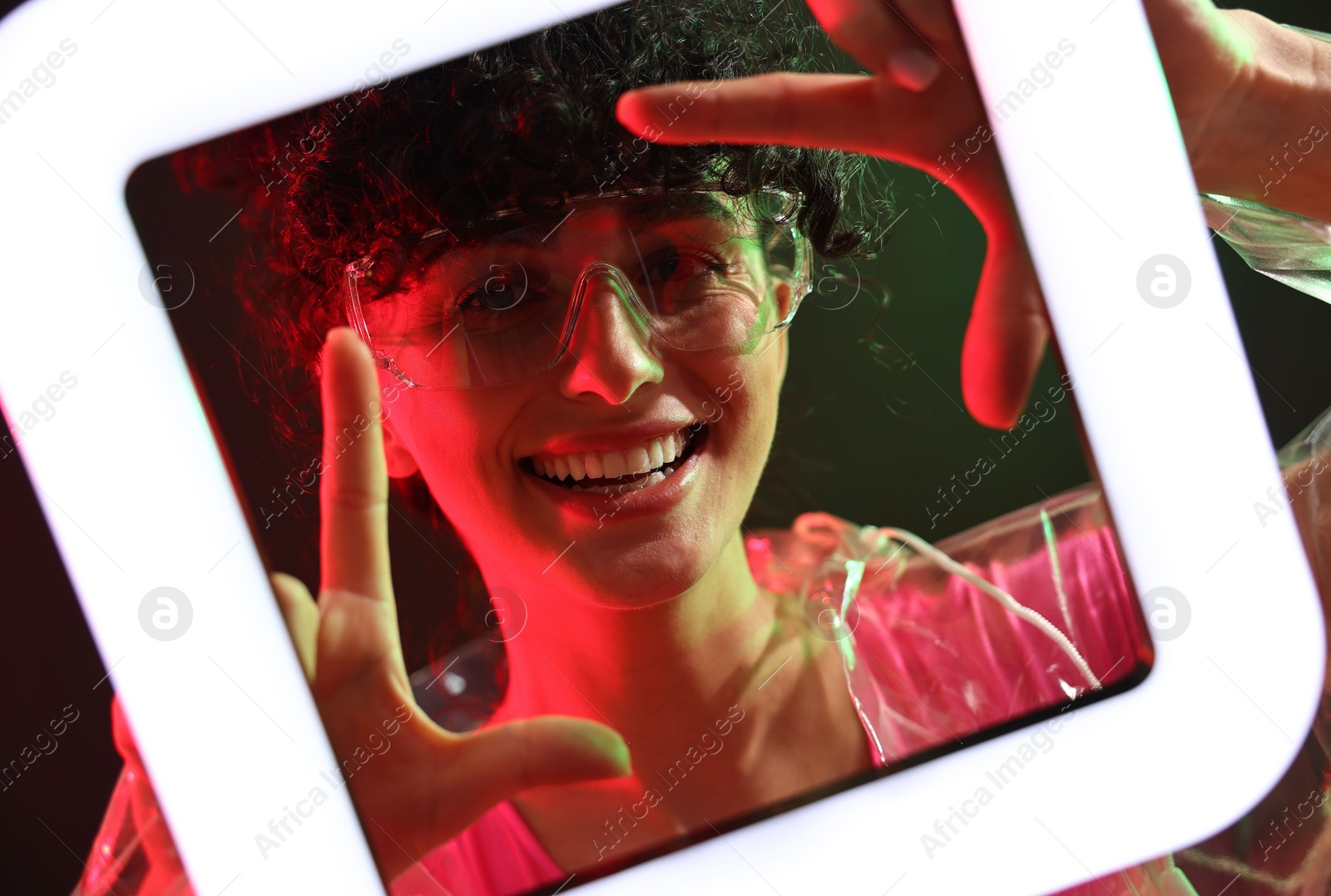 Photo of Beautiful young woman in transparent coat and sunglasses with square lamp on color background in neon lights