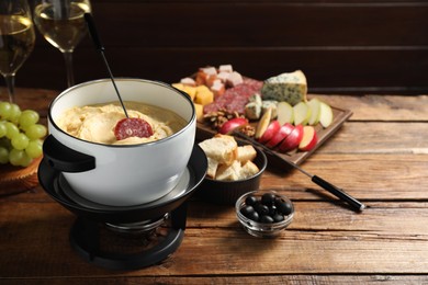 Photo of Fork with piece of sausage, melted cheese in fondue pot and other products on wooden table. Space for text