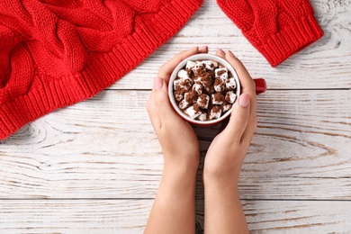 Photo of Woman holding cup of tasty cocoa with marshmallows on white wooden table, top view
