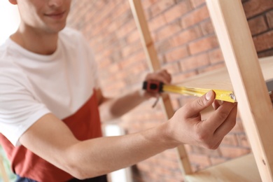 Photo of Young working man measuring wooden shelf indoors, closeup