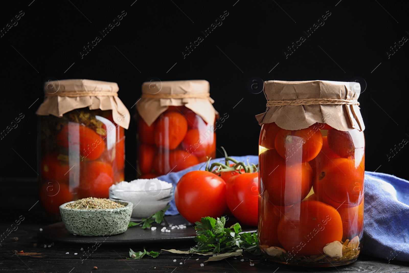 Photo of Pickled tomatoes in glass jars and products on black wooden table