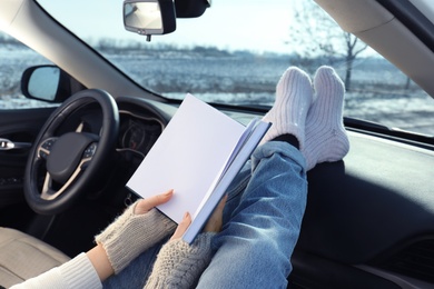 Photo of Young woman in warm socks holding her legs on car dashboard and reading book, closeup. Cozy atmosphere