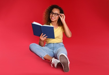 Beautiful African-American young woman reading book on red background